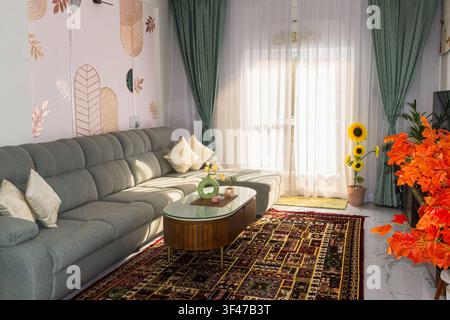 A stylish living room with a gray sectional, patterned rug, lush plants, and sunlight-filled windows creating warmth and modern elegance. Stock Photo