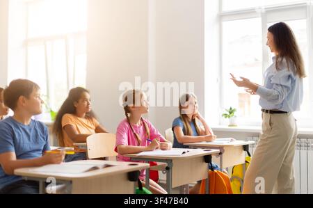 Female teacher explaining lesson to attentive children in bright multicultural classroom Stock Photo