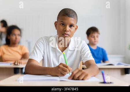 Serious black schoolboy writing in notebook at desk and looking toward camera Stock Photo