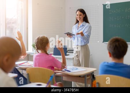 Smiling female teacher holding tablet engaging elementary students raising hands during lesson Stock Photo