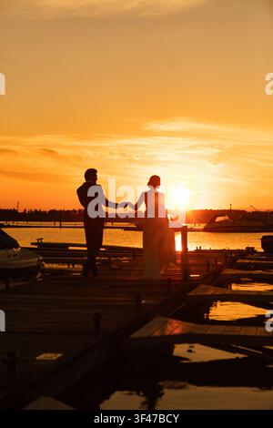 Bride and groom holding hands on marina pier during golden sunset Stock Photo