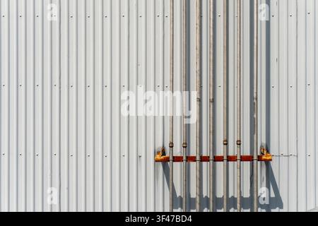 Multiple metal conduit pipes mounted on a white corrugated metal wall with a rusty bracket, creating geometric patterns and shadows in a minimalist in Stock Photo