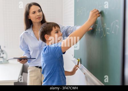 Focused schoolboy writing math equation on chalkboard while teacher holding tablet Stock Photo