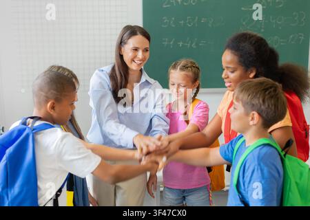 Female teacher and diverse schoolchildren stacking their hands together in classroom Stock Photo