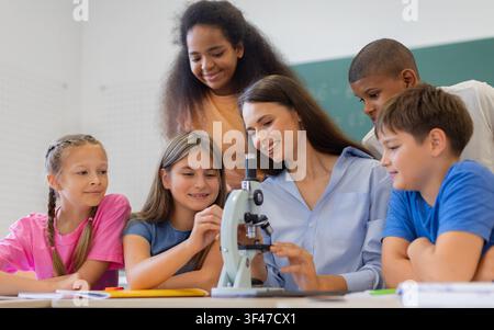 Diverse schoolchildren gathering around microscope with their teacher during practical science lesson Stock Photo
