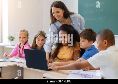 Female teacher assisting group of students using laptop at classroom desk Stock Photo