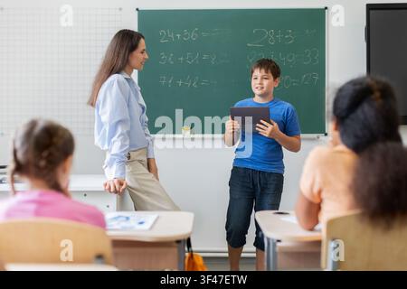 Smiling schoolboy holding tablet presenting math answer to teacher in classroom Stock Photo