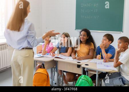 Female teacher explaining lesson to attentive diverse schoolchildren in classroom Stock Photo