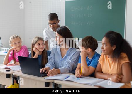 Female teacher using laptop while discussing classwork with diverse group of schoolchildren Stock Photo
