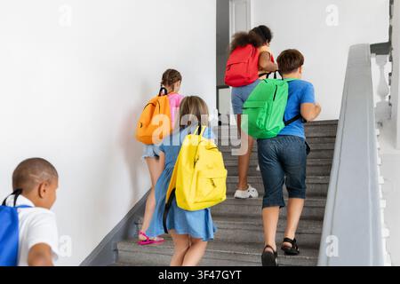 Diverse schoolchildren with colorful backpacks walking upstairs together inside school building Stock Photo