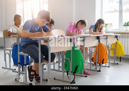 Diverse schoolchildren completing written assignments at individual desks in bright classroom Stock Photo