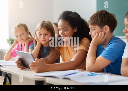 Diverse schoolchildren gathering around digital tablet in classroom Stock Photo