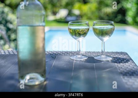 Bottle and two glasses of white wine on outdoor table near resort swimming pool. Stock Photo
