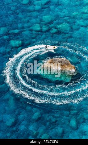 Aerial view of a small motorboat creating circular white waves around a rock in clear blue turquoise ocean water during summer vacation Stock Photo