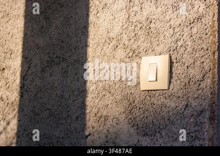 A close-up of a light switch on a textured wall. The wall has a rough surface with shadows cast by nearby objects. Stock Photo