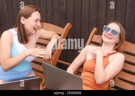 Two women relax on wooden deck chairs outdoors, laughing while using laptops. One wears sunglasses and a swimsuit, capturing a casual summer remote wo Stock Photo