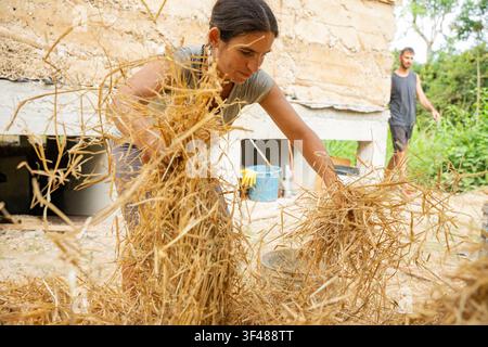 Woman preparing straw for a traditional bahareque house while another worker stands nearby during an ecological construction project using natural mat Stock Photo