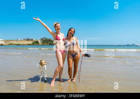 Two cheerful female friends in colorful bikinis pose and smile with a dog on a sandy beach in Ecuador, with calm waves, blue sky, and a coastal villag Stock Photo
