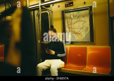 A solitary commuter sits on an orange bench inside a New York City subway car in Manhattan, looking at his smartphone beneath a transit map, urban tra Stock Photo