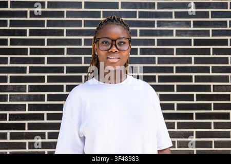 Smiling Black woman with long braids and eyeglasses wearing a white t-shirt, standing outdoors against a black-and-white brick wall, looking at the ca Stock Photo