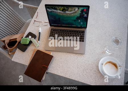Top view of a modern cafe workspace with laptop on a stone table, leather notebook, smartphone with earbuds case, pen, glasses, and a cup of coffee fo Stock Photo