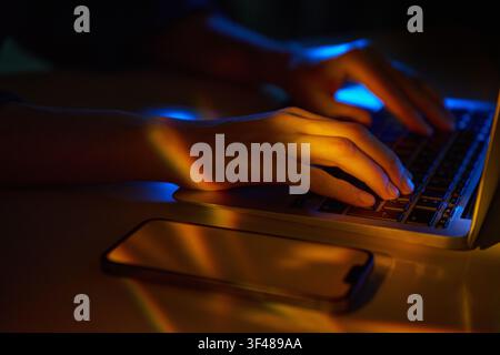 Close-up of hands typing on a laptop in neon light, with a smartphone on the desk. Futuristic hi-tech cyberspace mood for coding, hacking, and digital Stock Photo