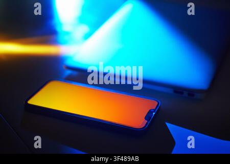 Close-up of a neon-lit smartphone beside an open laptop, glowing in hi-tech cyberspace. Vibrant blue and orange light creates a futuristic, digital at Stock Photo