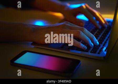 Close-up of hands typing on a hi-tech laptop beside a smartphone, illuminated by neon blue and magenta light, suggesting cyberspace security, coding, Stock Photo