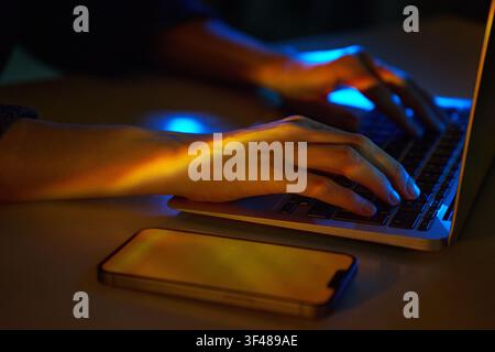 Close-up of hands typing on a laptop in neon light, with a smartphone on the desk. Hi-tech cyberspace mood for coding, hacking, network security, and Stock Photo