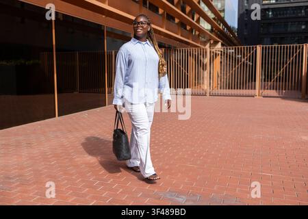 Confident Black woman with long braids walks through a modern city plaza, wearing business casual outfit and carrying a handbag, sunlight and contempo Stock Photo