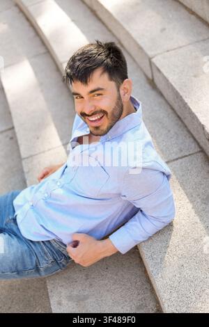 Smiling young man with beard in light blue shirt and jeans relaxes on sunlit stone steps outdoors, casual lifestyle portrait with warm daylight. Stock Photo