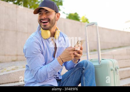 Happy young male traveler sitting on steps with rolling suitcase, wearing cap and headphones, checking smartphone and smiling in sunny urban setting. Stock Photo