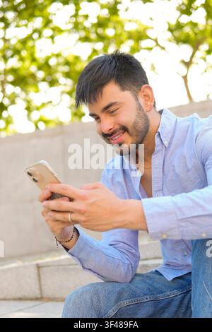 Smiling young man sitting on outdoor steps and texting on his smartphone. Casual lifestyle moment with natural light, green bokeh background, and rela Stock Photo