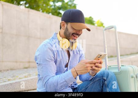 Smiling young man in cap with yellow headphones checks smartphone while sitting outdoors beside rolling suitcase, enjoying travel downtime, messaging Stock Photo