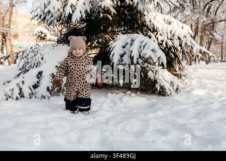 Warmly dressed toddler in a patterned snowsuit and knit hat stands in deep snow beside a snow-covered evergreen tree on a bright winter day in a city Stock Photo
