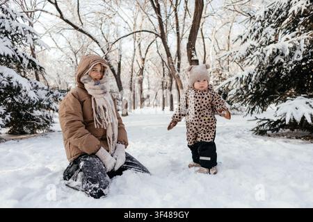 Smiling Girl in warm coat and scarf sits in fresh snow while her toddler in a patterned snowsuit and hat stands nearby on a snowy forest path in winte Stock Photo