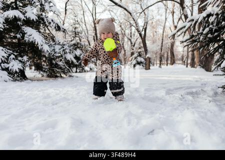 Warmly dressed toddler stands in a snowy park, holding a bright toy shovel and bucket, enjoying outdoor winter play among snow-covered trees on a cold Stock Photo
