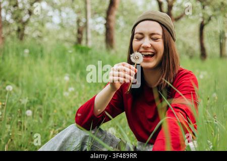 Happy young woman in red sweater and beanie sits in a green spring meadow, laughing while holding a dandelion seed head, surrounded by grass and trees Stock Photo