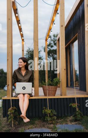 Young woman sitting on tiny house patio, working on laptop. Stock Photo