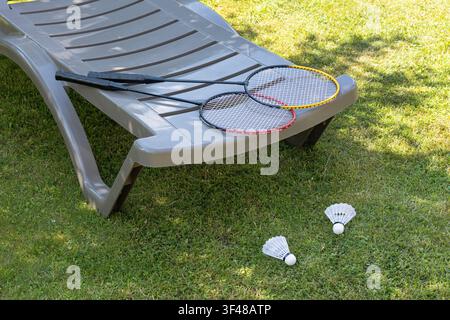 Two badminton rackets resting on a gray garden sun lounger with shuttlecocks on the grass, capturing a sunny backyard leisure scene and outdoor sport Stock Photo
