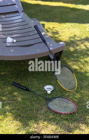 Two badminton rackets and shuttlecocks rest on green grass beside a sunlit patio lounge chair, suggesting outdoor summer recreation, leisure, and back Stock Photo