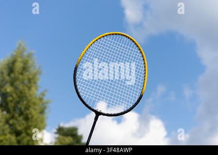 Close-up of a badminton racket held up against a bright blue sky with soft white clouds, outdoor sport concept and summer leisure atmosphere. Stock Photo