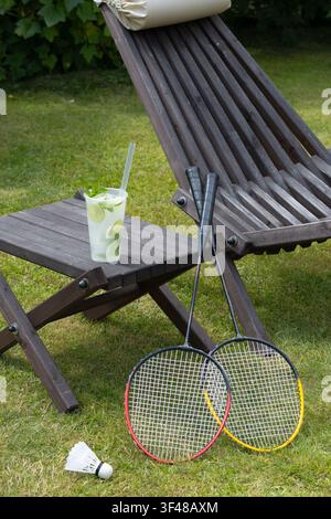 Summer garden scene with wooden deck chair, folding side table and refreshing lemonade with mint. Two badminton rackets and a shuttlecock on green law Stock Photo