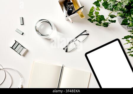Bright minimalist desk flat lay with tablet mockup, glasses, notebook and pen, stapler, ruler and green plant on a clean white background. Stock Photo