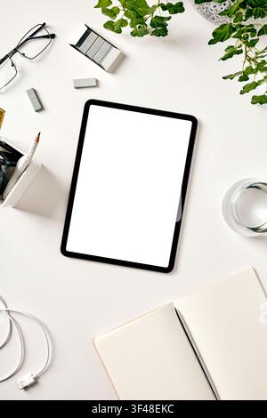 Top view of a clean white workspace with a blank tablet screen mockup, glasses, notebook, pen, pencil holder, USB cable, staples, water glass, and gre Stock Photo