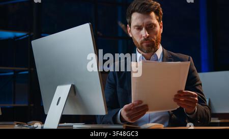 Concentrated Caucasian middle-aged business man analyzing papers with computer in night office focused businessman male work with documentation legal Stock Photo