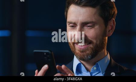 Caucasian happy optimistic mature business man smile using chatting mobile phone at night office positive joyful businessman executive male with Stock Photo