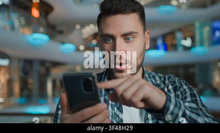 Happy Caucasian man scrolling mobile phone app excited amazed pointing in screen in shopping mall positive business male client businessman guy with Stock Photo