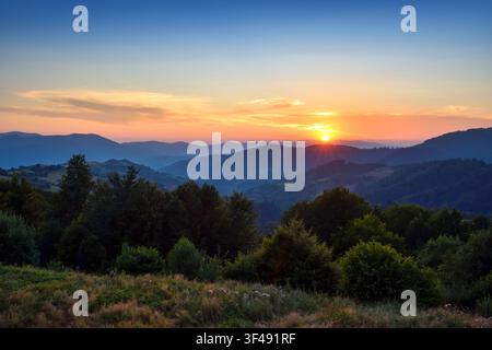 Beautiful sunset over mountain landscape with forest, rolling hills and colorful sky. Peaceful summer nature panorama. Stock Photo
