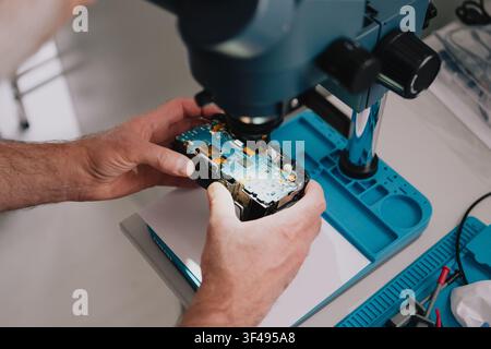Electronics technician inspecting a disassembled digital camera circuit board under a stereo microscope at a repair workbench Stock Photo